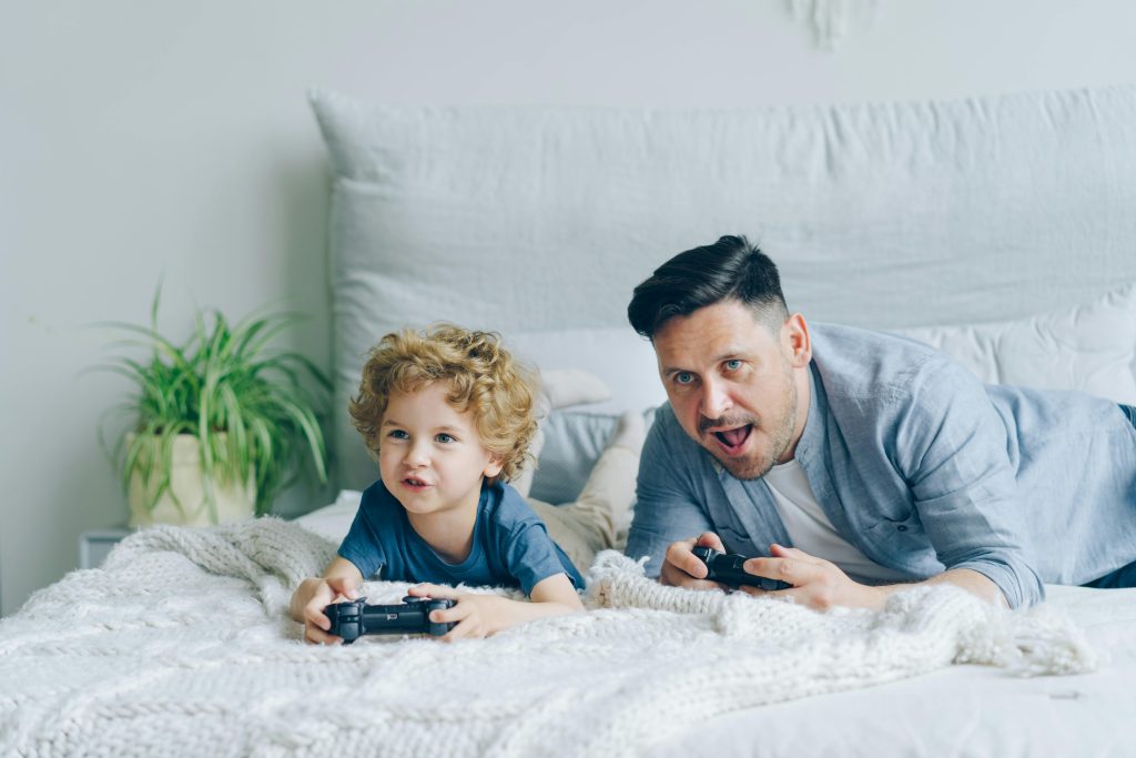 Man and His Little Son Lying on a Bed and Playing a Video Game